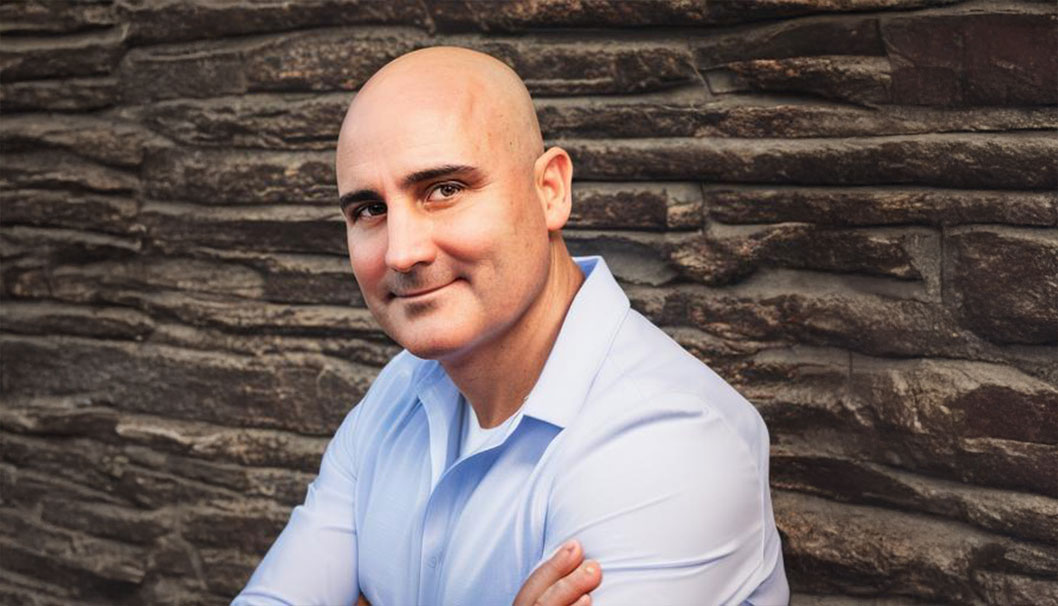 A bald man in a blue shirt leans against a wall